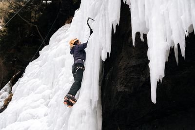 Person with helmet and crampons climbs with ice axe on icy rock face in a mountainous environment.
