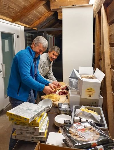 Two people are preparing food on a table with packaging in a room with wooden walls.