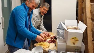 Two people are preparing food on a table with packaging in a room with wooden walls.
