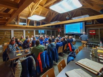 A group of people is sitting in a wooden room at tables while a presentation is being shown.
