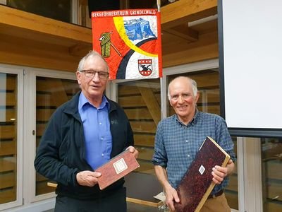 Two men are holding books in front of a banner of the Mountain Guide Association Grindelwald in a wooden building.