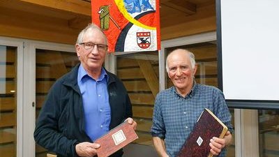 Two men are holding books in front of a banner of the Mountain Guide Association Grindelwald in a wooden building.