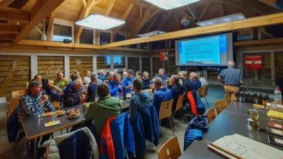 People are sitting at tables in a room with a wooden roof, a man is standing in front of a projector screen.