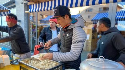 People are serving food at a market stall with blue and white awnings in the background.