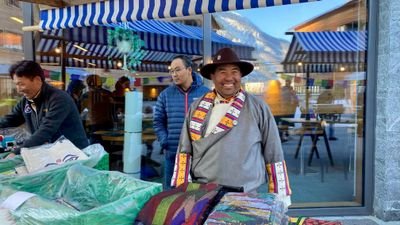 People at a market stall with colorful textiles, with mountains and buildings in the background.