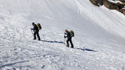Two people with backpacks and sticks are hiking in the snow in a mountainous landscape.