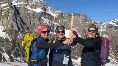 Three people with backpacks and sunglasses are holding an ice axe in front of snow-covered mountains.