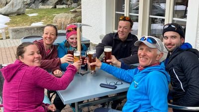Six people in outdoor clothing clink beer glasses at a table outdoors.