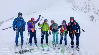 Six people with skis and climbing equipment are standing on a snow-covered mountain peak.