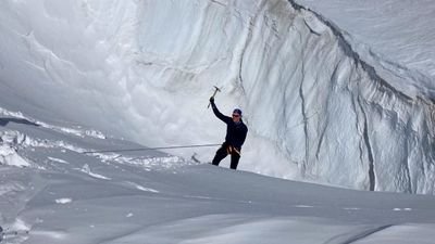 Person with helmet and climbing gear on a snowy glacier, holding an ice axe, surrounded by ice walls.