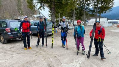 A group of five people with skis and ski equipment is standing in a parking lot in the mountains.