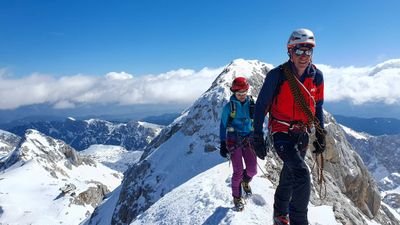 Two people with helmets and climbing gear on a snow-covered mountain ridge in an alpine environment.