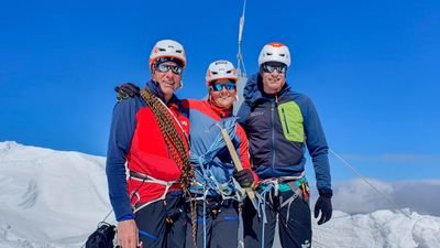 Three people with helmets and climbing gear are standing on a snow-covered mountain peak.