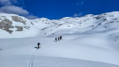 People with skis and poles are walking in snowy mountains. Clear sky in the background.