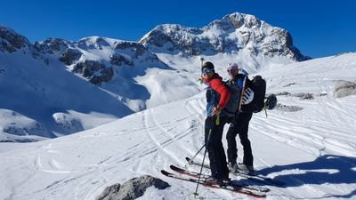 Two people with skis and helmets are standing on a snowy mountain in front of an alpine backdrop.