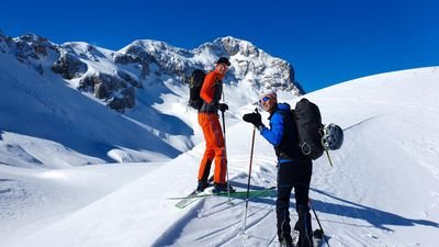 Two people with skis and backpacks on a snowy mountain, wearing helmets and ski poles.