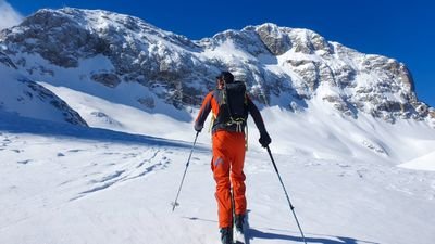 Person with skis and backpack in snowy mountain landscape, blue sky in the background.