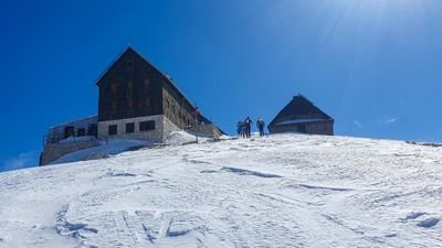 People with skis and poles in front of a mountain hut in the snow, blue sky in the background.