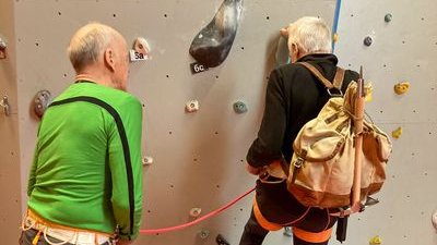 Two people are climbing in a hall with climbing harnesses and ropes on a climbing wall.