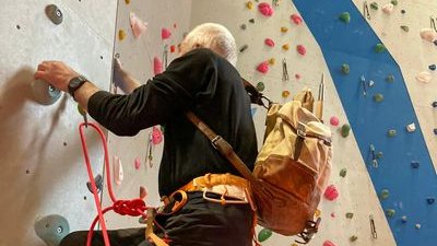 Person climbing in a hall with a climbing harness and rope, colorful holds on the wall.