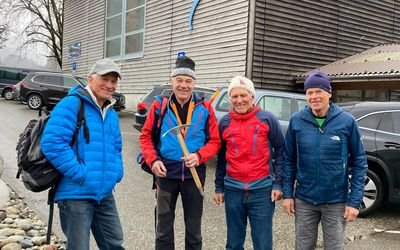 Four people in outdoor clothing and hats are standing in front of a building with cars in the background.