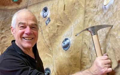 A person is holding an ice pick in front of a climbing wall in an indoor climbing gym.