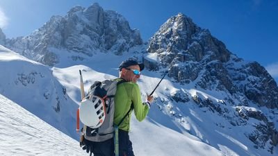 Person with helmet and backpack on snow-covered mountain, holding a hiking pole; high peaks in the background.