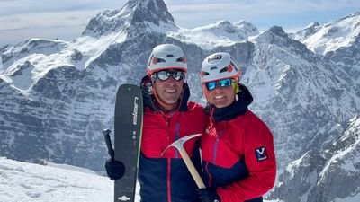 Two people in red jackets with helmets and ice axes in front of snow-covered mountains.
