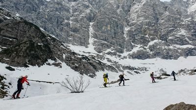 People with skis and backpacks are climbing a snowy mountain, surrounded by rocky peaks.