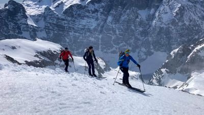 Three people with skis and poles are skiing on a snowy mountain, surrounded by high peaks.