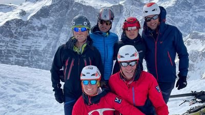 Six people with helmets and ski goggles are standing in snowy mountains.