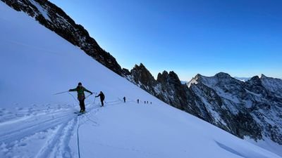 People with skis and poles on a snowy mountain, secured with a rope, surrounded by peaks.