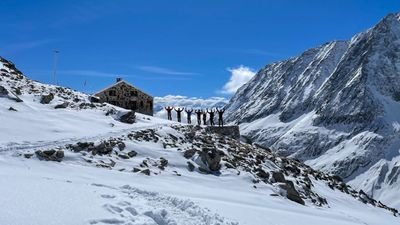 People are standing in a snowy mountain landscape in front of a hut, surrounded by high peaks.