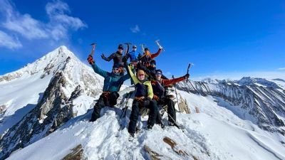 Group of people with ice picks on a snow-covered mountain peak, surrounded by an alpine landscape.