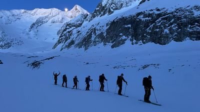 Group of people with skis and backpacks in the snowy mountains at dusk.