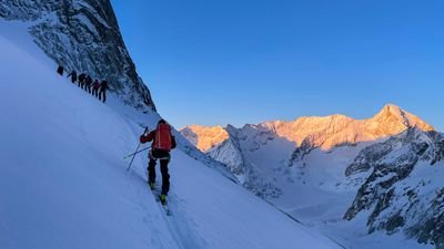 People with ski equipment are ascending a snow-covered slope in a mountain range at sunrise.