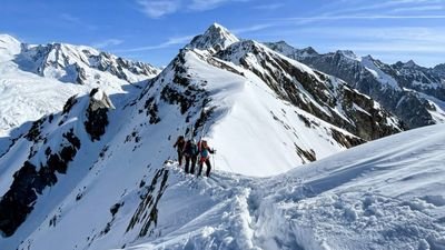 Three people with helmets and climbing gear on a snow-covered mountain ridge in the Alps.