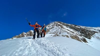 People with helmets and climbing gear are hiking on a snow-covered mountain path under a blue sky.
