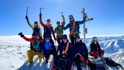 Group of mountaineers with helmets and ice axes on a snowy summit in front of a mountain panorama.