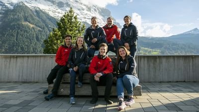 Group of people in outdoor clothing sitting on a bench in front of a mountain scenery.