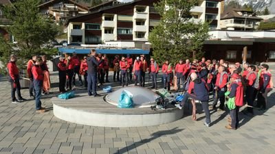 Group of people in red jackets gathered outdoors in front of buildings in a mountainous region.