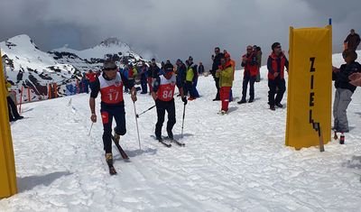Two skiers with race numbers and poles pass a finish banner in the snowy mountains.