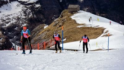 Three people with race numbers and skis are climbing a snow-covered slope in the mountains.