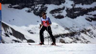 Person on skis with poles on a snow-covered mountain, wearing a vest with a number, with rocks in the background.