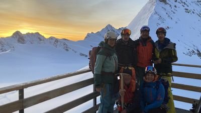 Group with helmets and winter clothing on wooden platform in front of snow-covered mountains at sunset.