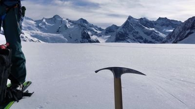 Person with skis and ice pick on snowy glacier in front of mountain scenery.