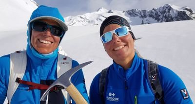 Two people in blue outdoor clothing with sunglasses and ice axes in snow-covered mountains.