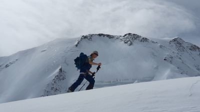Person with helmet and backpack climbs a snow-covered mountain with ski poles.