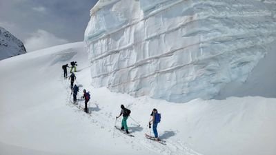 People with skis and backpacks are climbing in a row up a snowy mountain next to an ice wall.