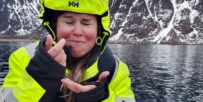 A person in a yellow jacket on a boat in front of snow-covered mountains and water.